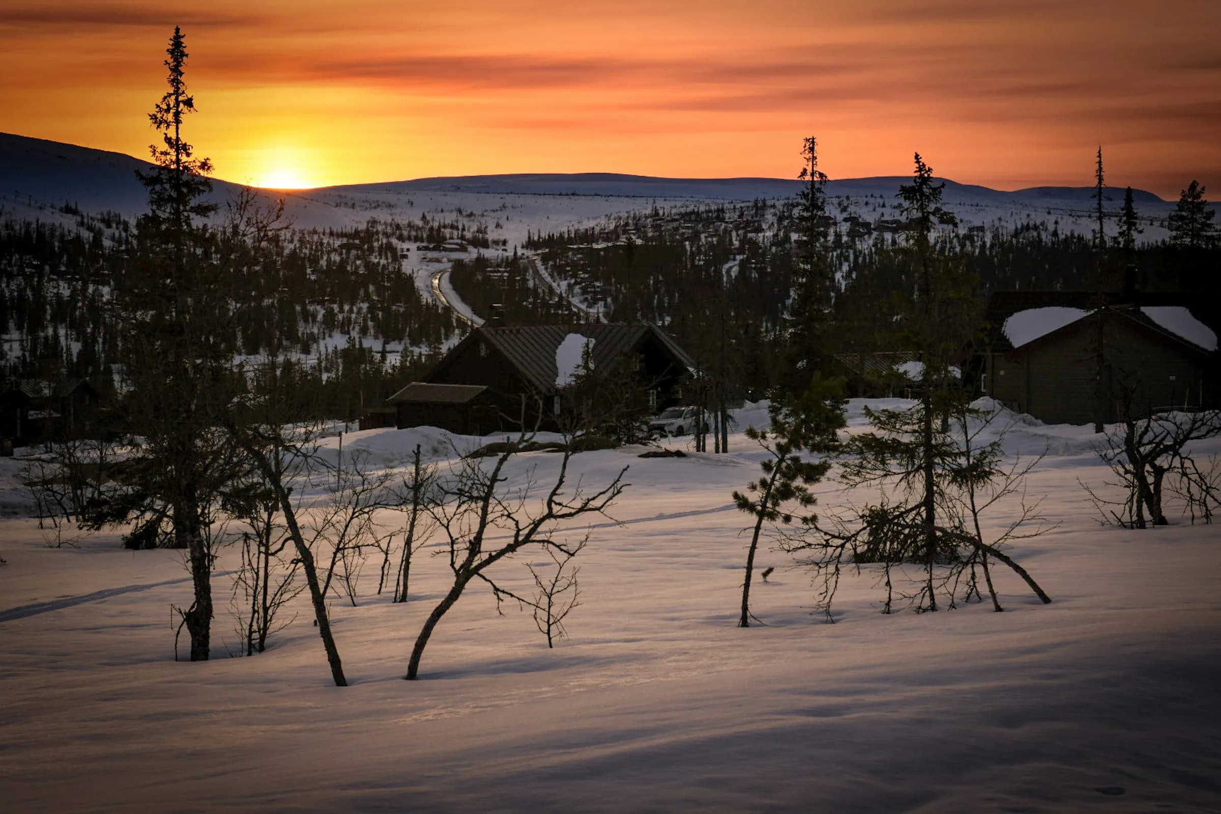 Solnedgång över snötäckta hus och glesa träd i fjällterräng.