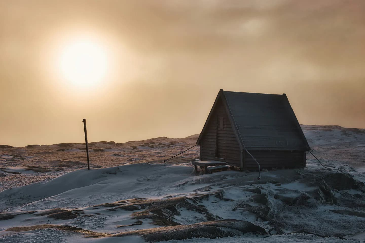 Liten stuga på en snötäckt fjällhöjd med solen genom dimman.